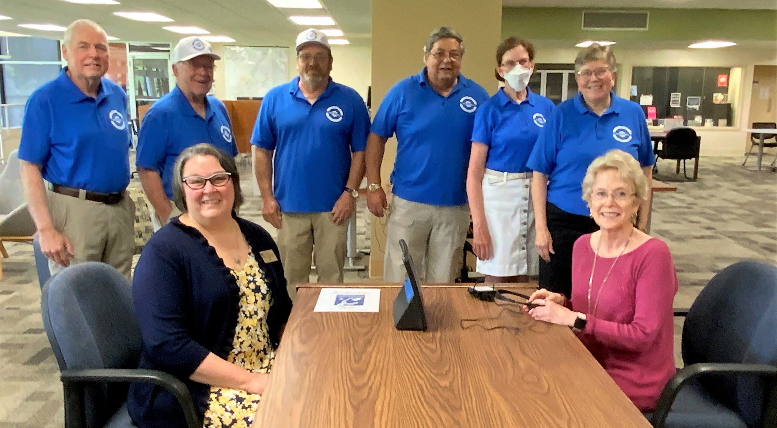 Tammy Kuhn-Schnell and Dr. Charlotte Warren seated at a table with the hearing loop device, and members of the Sertoma Club standing behind