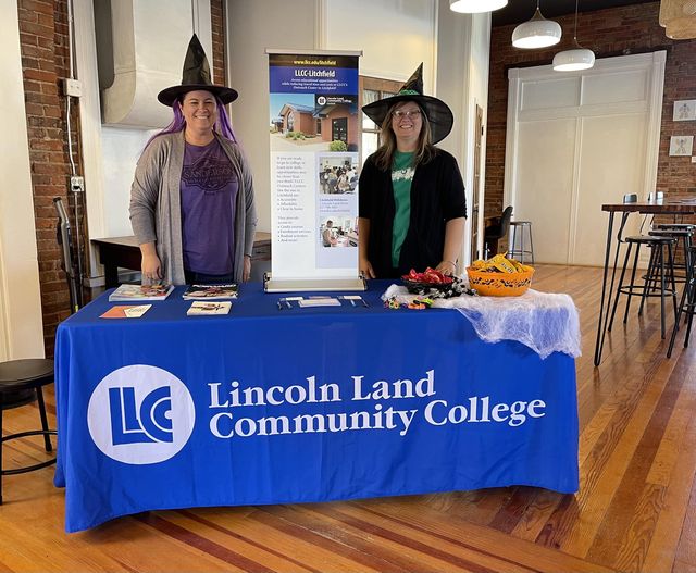 Jessie Blackburn and Tisha Miller wearing witches hats and standing at an LLCC info table