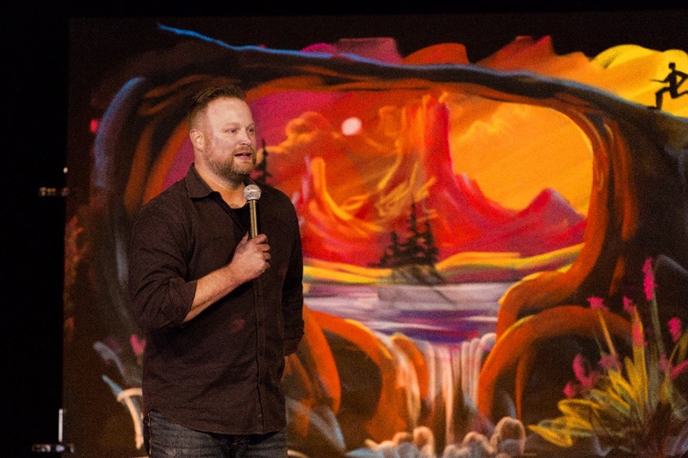 Photo of the Chalkguy speaking on a microphone in front of his artwork