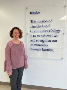 Anne Armbruster standing next to a sign with the college's mission. The mission of Lincoln Land Community College is to transform lives and strengthen our communities through learning.