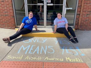 Two students by LLCC-Jacksonville doors and sidewalk that says "Only yes means yes. Sexual Assault Awareness Month."