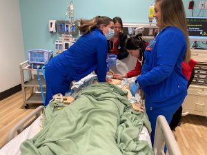 Nursing student providing chest compressions with other students around patient bed in sim lab