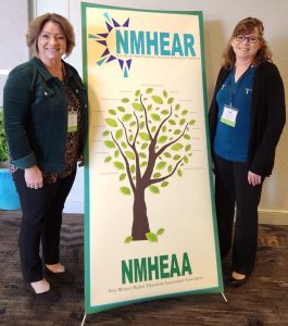 Lisa Avendano and Laurie Myers at conference, standing on either side of an NMHEAR/NMHEAA sign.