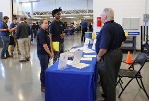 Exhibit area in background. Two students talking to employer in foreground.