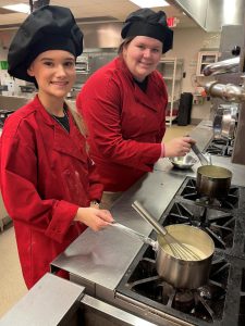 Two students in red chef coats cooking on a stove