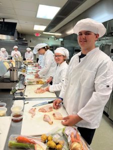 Many students in white chef coats and hats standing around prep tables, preparing chicken