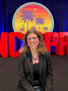 Kyla Kruse sitting on a stage with the letters NCMPR behind her and a logo with palm trees