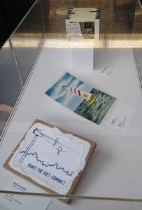 Glass case containing three pieces of artwork. The first piece is an illustration done in blue marker on white paper with the words “How’s the diet coming?” on it. It has one thin bird sitting on a regular telephone wire, and a larger bird sitting on a sagging telephone wire. Another piece of artwork is a painting of a yellow sailboat with a red striped sail that has the sun on it, sailing on deep green/blue water with a blue and white sky behind it. Third piece of artwork is a paper accordion fold piece with Abraham Lincoln on the front with the words “The day Lincoln was shot.”