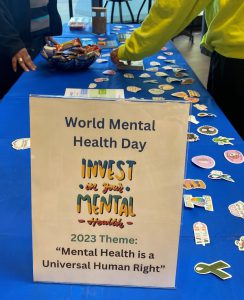 Table with people picking up stickers and snacks. Sign that says World Mental Health Day. Invest in your mental health. 2023 theme: "Mental Health is a Universal Human Right."