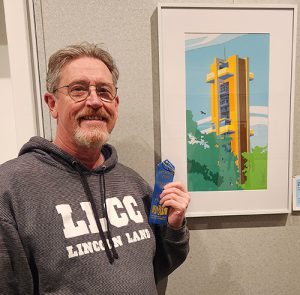 Greg Walbert holding a blue ribbon, standing in front of his “Hear the Bells Ringing” artwork which is an image of a carillon behind trees made out of a variety of paper.