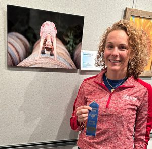 Michelle Burger holding a blue ribbon, standing in front of her “Bale Yeah Cowgirl” artwork which is a photograph of a young girl doing a backbend on a bale of hay.