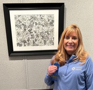 Diane M. Wilson holding a blue ribbon, standing in front of her “Flower Fantasy II” artwork which is a B/W drawing of a variety of flowers and leaves.