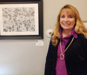Diane M. Wilson standing next to her white third-place ribbon and “Flower Fantasy II,” which is a B/W drawing of a variety of flowers and leaves.