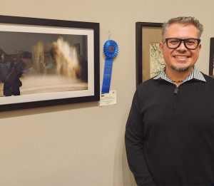 Esteban Cruz standing next to his blue first-place ribbon and “Rain, Rain, Go Away,” which is a photograph at night of the rain with a shadow of a person in the foreground.