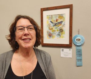 Jan Szoke standing next to her light blue Honorable Mention ribbon and “Summer’s Glory,” which is a mixed media artwork of flowers, child, bird nest and a bird made with ink, watercolor, paper.