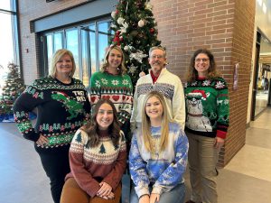 Public Relations and Marketing staff wearing ugly holiday sweaters and standing in front of decorated tree