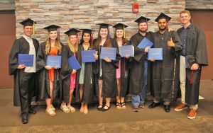 Group of nine graduates in cap and gown, holding diplomas.