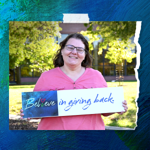 Lady in a pink shirt smiling. Holding a sign that reads "Believe in giving back."