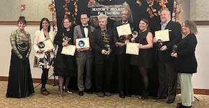 The photo shows nine people dressed in formal attire standing in front of an autumn-themed backdrop that says "Mayor's Awards for the Arts."  The woman on the far left designed the stained glass awards, which the eight award winners hold, along with their certificates.